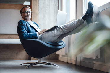 Senior male entrepreneur sitting with laptop on lap at office - GUSF05788