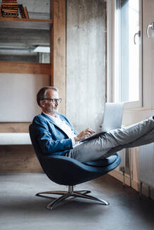 Männlicher Unternehmer mit Laptop auf dem Schoß, während er im Büro sitzt - GUSF05789