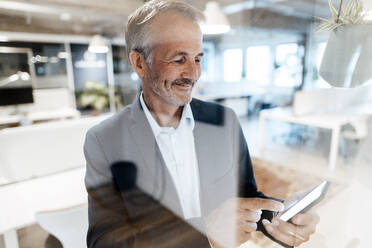 Senior businessman using mobile phone by glass in office - GUSF05790