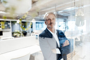 Senior male entrepreneur with arms crossed by glass wall in office - GUSF05791