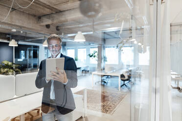 Ein älterer Geschäftsmann benutzt ein digitales Tablet, während er hinter einer Glaswand steht - GUSF05798