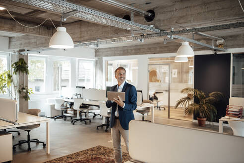 Lächelnder Geschäftsmann, der ein digitales Tablet hält, während er im Büro steht - GUSF05814