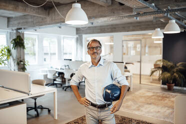 Älterer männlicher Architekt, der einen Schutzhelm hält, während er mit der Hand auf der Hüfte im Büro steht - GUSF05816