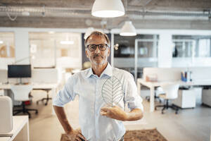 Confident senior businessman holding leaf model with hand on hip at office - GUSF05818