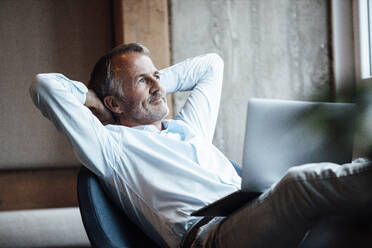 Thoughtful businessman looking away while sitting with laptop in office - GUSF05833