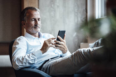 Älterer männlicher Unternehmer, der im Büro sitzend ein Mobiltelefon benutzt - GUSF05837