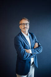 Confident businessman standing with arms crossed in front of blue wall - GUSF05842