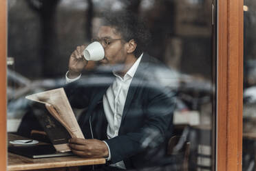 Businessman drinking coffee while reading newspaper sitting in cafe seen through glass - GUSF05862