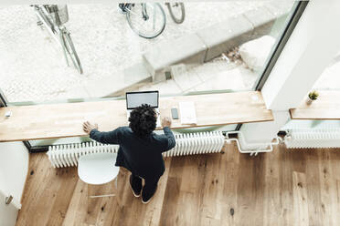 Male entrepreneur with laptop standing by desk looking through window in office - GUSF05863