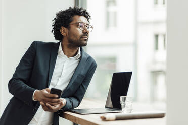 Nachdenklicher männlicher Unternehmer mit Smartphone, der sich auf den Schreibtisch stützt und durch ein Fenster im Büro schaut - GUSF05876