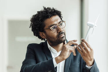 Männlicher Unternehmer mit lockigem Haar, der ein Windradmodell im Büro untersucht - GUSF05878