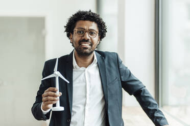 Smiling mature businesswoman standing with wind turbine model in office - GUSF05880