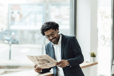 Lächelnder männlicher Unternehmer, der Zeitung liest, während er sich in einem Büro am Fenster anlehnt - GUSF05884