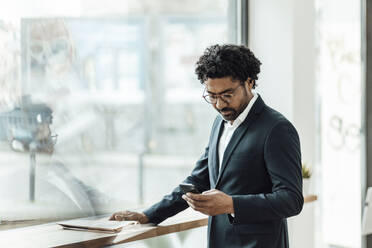 Männlicher Unternehmer, der ein Smartphone benutzt, während er an einem Glasfenster im Büro steht - GUSF05887