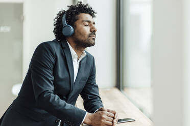 Curly haired businessman with eyes closed listening music through headphones while leaning on desk in office - GUSF05893