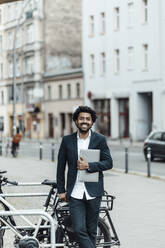 Lächelnder reifer Geschäftsmann, der einen Laptop hält und sich auf ein Fahrrad in der Stadt stützt - GUSF05900