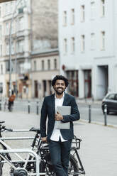 Smiling mature businessman holding laptop while leaning on bicycle in city - GUSF05900