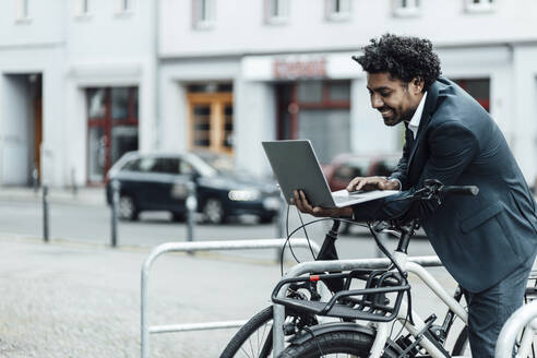 Lächelnder Geschäftsmann, der einen Laptop benutzt, während er sich auf einem Fahrrad in einer Parkstation in der Stadt abstützt - GUSF05903