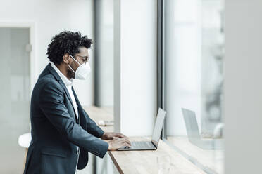 Mature businessman wearing protective face mask using laptop in office during COVID-19 - GUSF05904