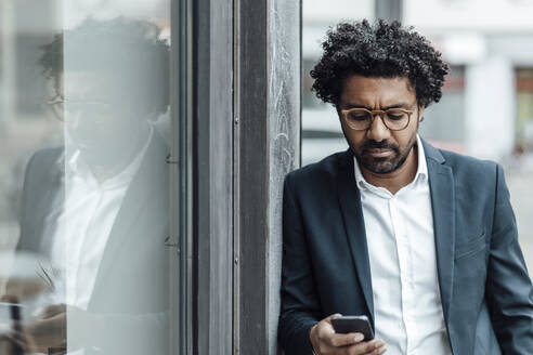 Männlicher Berufstätiger, der an der Wand lehnt und mit seinem Mobiltelefon SMS schreibt - GUSF05910