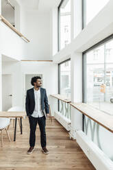 Smiling male entrepreneur standing with laptop in office looking through window - GUSF05911