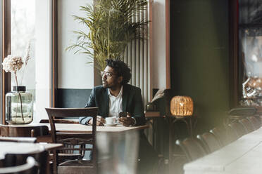 Älterer Geschäftsmann mit Mobiltelefon sitzt mit Kaffee im Café und schaut weg - GUSF05914