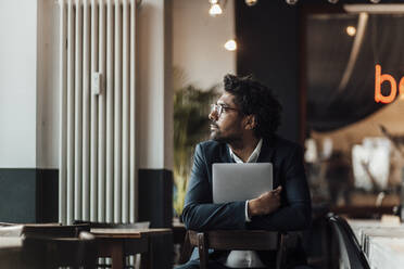 Male entrepreneur holding laptop while sitting on chair in cafe looking away - GUSF05921