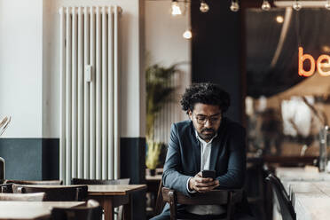 Reifer männlicher Berufstätiger, der mit seinem Smartphone im Internet surft, während er auf einem Stuhl im Café sitzt - GUSF05922