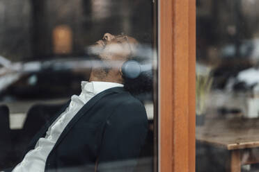 Männlicher Berufstätiger mit geschlossenen Augen hört Musik über Kopfhörer in einem Café, gesehen durch Glas - GUSF05926