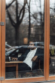 Geschäftsmann, der einen Laptop benutzt, während er im Café Musik hört, gesehen durch Glas - GUSF05927