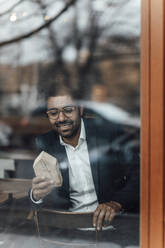 Lächelnder männlicher Unternehmer, der ein Hausmodell betrachtet, während er in einem Café sitzt, gesehen durch Glas - GUSF05930