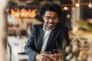 Lächelnde männliche Fachkraft, die in einem Café eine Textnachricht über ihr Smartphone verschickt - GUSF05940