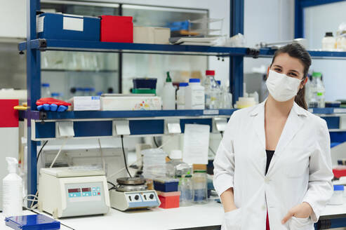 Junge medizinische Forscherin mit den Händen in den Taschen im Labor während einer Pandemie - PGF00589