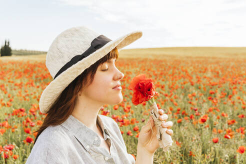 Schöne Frau mit Hut riecht an Mohnblumen auf einem landwirtschaftlichen Feld - MGRF00243