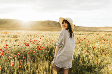 Smiling woman wearing hat and shirt standing at poppy field during sunny day - MGRF00253