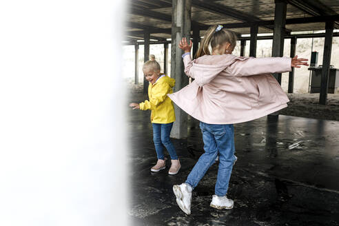 Sisters wearing raincoats playing under roof at cafe - OGF01049