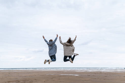 Carefree female friends jumping with arms raised at beach - JMPF00962