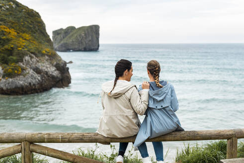 Female friends sitting on wooden fence in front of sea - JMPF00969