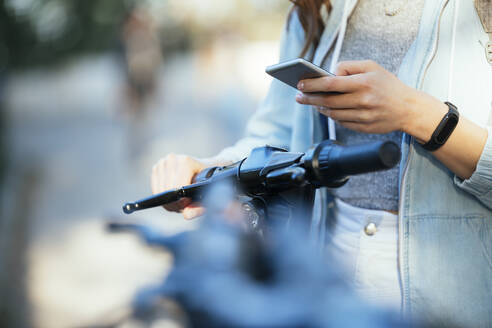 Frau, die eine Armbanduhr trägt, benutzt ein Mobiltelefon über einem Motorroller - OYF00377