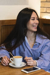 Lächelnde junge Frau mit Kaffeetasse, die wegschaut, während sie in einer Bar sitzt - PNAF01682