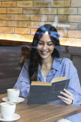 Smiling young woman reading book while sitting in bar seen through glass - PNAF01685