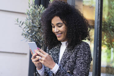 Smiling woman using smart phone near window outside bar - PNAF01689