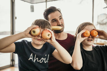 Verspielte Brüder bedecken ihre Augen mit Früchten, während sie mit ihrem Vater in der Küche sitzen - MJRF00576