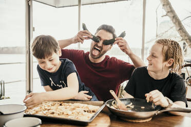 Verspielter Vater spielt mit seinen Söhnen, während er in der Küche eines Hausbootes Essen zubereitet - MJRF00578