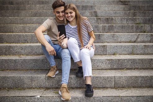 Lächelndes Paar, das ein Selfie mit dem Mobiltelefon macht, während es auf den Stufen sitzt - GRCF00734