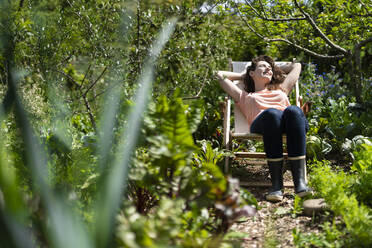 Junge Frau entspannt sich auf einem Stuhl im Garten an einem sonnigen Tag - SBOF03789
