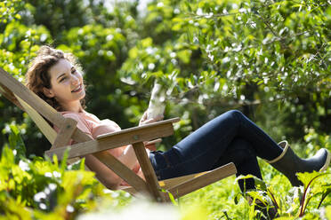 Junge Frau schaut weg, hält ein Buch und sitzt auf einem Stuhl im Garten - SBOF03791