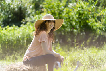 Frau mit Sonnenhut sitzt auf Heu im Garten an einem sonnigen Tag - SBOF03794