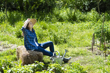 Frau sitzt auf einem Stapel Heu im Permakultur-Garten an einem sonnigen Tag - SBOF03795