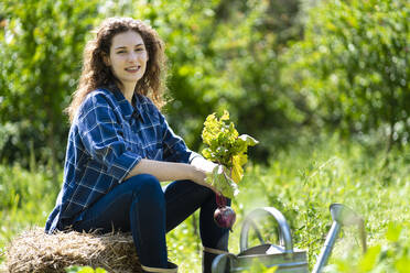 Lächelnde junge Frau sitzt auf einem Stapel Heu und hält eine Rote-Bete-Pflanze - SBOF03798